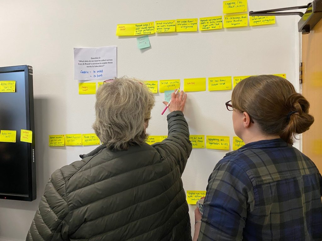 A group of two Energise Manchester team members stand in front of a wall with lots of sticky notes listing retrofit measures. One of them is writing on a sticky note.
