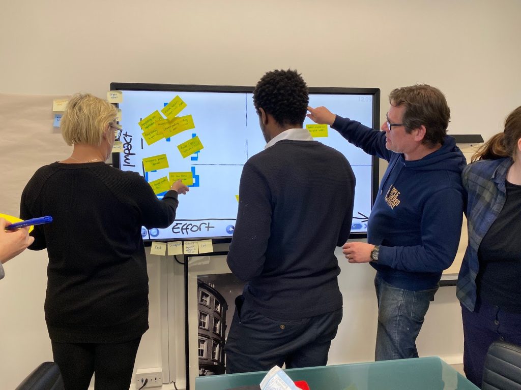 A group of three Energise Manchester team members stand in front of an interactive whiteboard with a chart that has Impact on the Y axis and Effort on the X axis. They’re putting sticky notes with retrofit measures on them onto the board.

