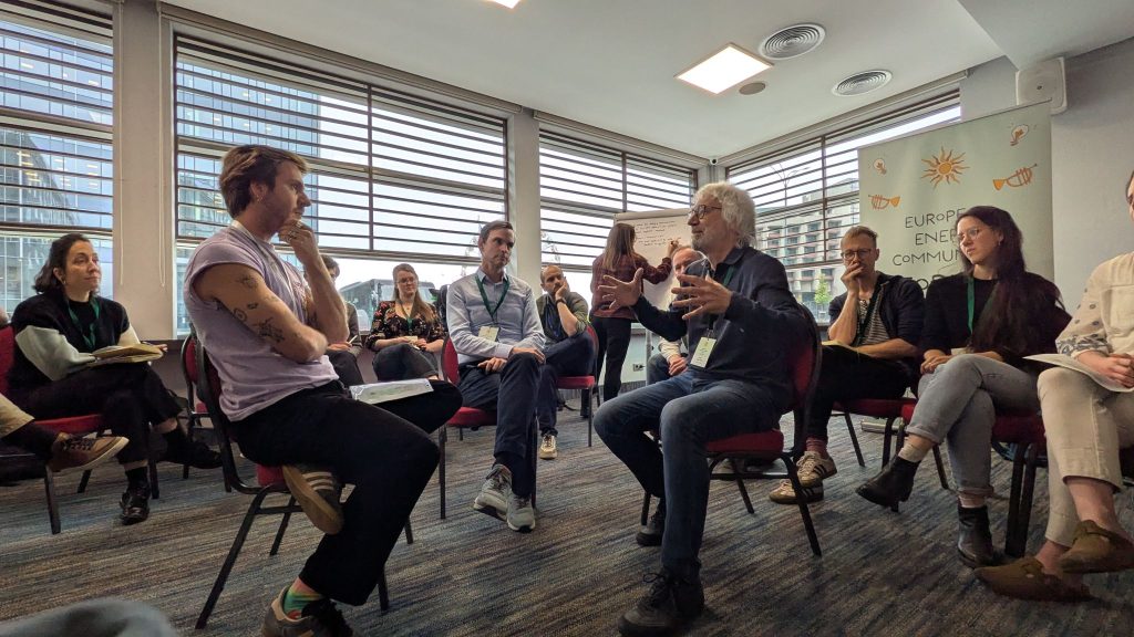 A group of people are seated in a circle for a discussion in a brightly-lit conference room. In the foreground, an older man with grey hair and glasses gestures with his hands as he speaks to a younger man in a purple t-shirt, who is listening intently. Other participants are seated in the background, and a banner for the "European Energy Community" is partially visible on the right.